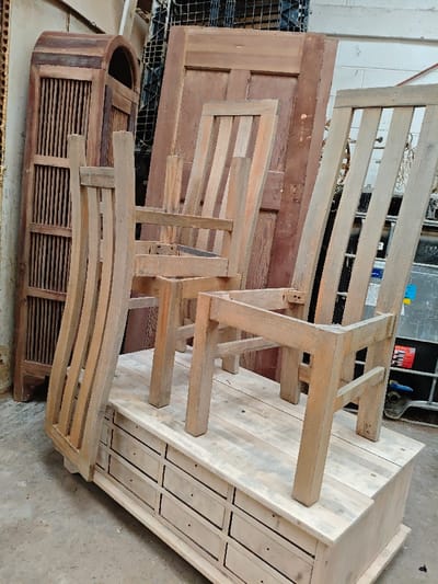 The image displays an indoor storage or workshop area in Swindon, United Kingdom. In the foreground, a low, light-colored wooden cabinet or chest of drawers with multiple small, unfinished drawer fronts is positioned on a concrete floor. Three wooden chair frames rest on top of this cabinet. One chair frame, made of light wood with a curved backrest and vertical slats, is positioned on its side, leaning left. Two other light-colored wooden chair frames stand upright, featuring straight backrests with vertical slats and exposed joinery, indicating they are incomplete or disassembled. In the background, a tall, dark reddish-brown wooden cabinet with an arched top and a slatted front panel stands against a wall. Adjacent to it, a large, dark reddish-brown wooden door leans vertically. Further background elements include metallic grating, a dark industrial container with visible labels, and a partial white wall. No individuals or active interactions are present in the scene.