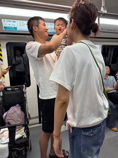 The image depicts an indoor scene, likely a subway car, with a family of three. A man, wearing a white t-shirt and black shorts, is holding a distressed young child who is wearing a striped shirt. The child appears to be crying or yelling. A woman, seen from behind and the side, is wearing a white t-shirt and blue jeans with a green strap across her shoulder. She has her hair pulled up in a bun. They are standing near a black stroller with various items inside, including a pink bag with the label "onnichoco" and some fabric. In the background, other passengers are visible, including a man looking at his phone and another man reflected in a window. A digital display board above the man and child shows information about the Guangzhou Metro, specifically indicating that the next station is Chigang. This suggests the location is a subway in Guangzhou, China, likely within the Baiyun District given the context of the metro system.