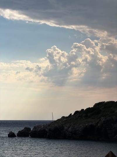 This media file is a photograph of a dramatic sky and seascape. The image is oriented vertically, with the sky occupying the top two-thirds and the sea and a rocky cliff face the bottom third.
The sky is filled with a dynamic display of clouds. On the left, a large, dark, and striated cloud formation stretches vertically, appearing almost like a storm front. To its right, fluffy, white cumulus clouds are illuminated by sunlight, creating a contrast between light and shadow. Rays of light are visible breaking through the clouds in the center of the frame. The sky transitions to a lighter, hazy hue towards the right horizon, suggesting a sunset or sunrise.
The bottom of the image features the dark, textured surface of a rocky cliff face on the right side. Below the cliff is the dark, rippling surface of the sea, reflecting the soft light of the sky. Three dark, irregularly shaped rocks are partially submerged in the sea in the foreground. A thin, dark mast of a sailboat is visible against the sky, just above the cliff line, hinting at a distant vessel.
The time of day appears to be either early morning or late evening, given the soft, diffused light and the hazy horizon. The weather seems to be mixed, with clear sky and bright sunlight interspersed with darker cloud formations.
The overall mood is serene and somewhat dramatic, evoked by the contrast between the vast sky and the rugged coastline. There is no visible text in the image.