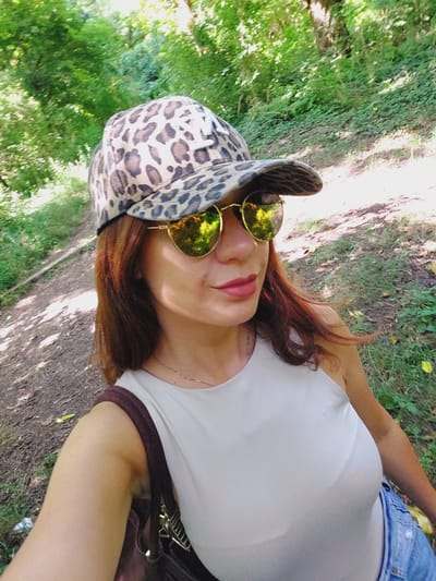 In Tuchenitsa, Bulgaria, a woman with auburn hair is pictured from the chest up, wearing a leopard print baseball cap and round, gold-rimmed sunglasses. She is dressed in a light-colored sleeveless top and blue jeans. The background features a wooded area with a dirt path and green foliage. She is holding a dark-colored bag on her left shoulder.