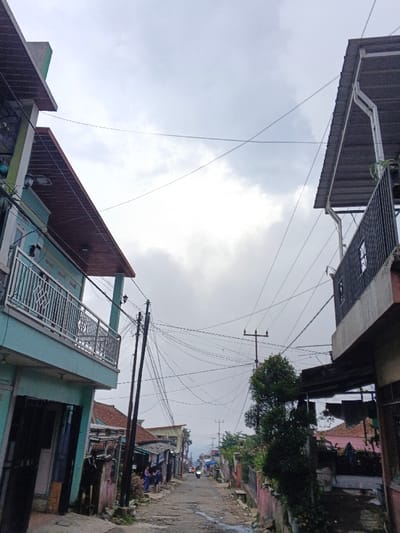The image displays a narrow, sloping residential street under an overcast sky. Two-story buildings line both sides of the thoroughfare. On the left, a light teal-colored building features a second-story balcony with a silver-toned metal railing adorned with a diamond pattern, beneath a brown-colored roof overhang. Its ground level presents dark, recessed openings. On the right, a building with a dark grey corrugated metal roof exhibits a white rain gutter and a dark metal balcony railing; a small green tree partially obscures its lower facade.

The street surface is uneven, consisting of unpaved ground with visible dirt and depressions. Multiple utility poles stand along the street, supporting a dense network of black overhead utility wires that crisscross the frame against the pale, cloudy sky.

Further down the street on the left, two individuals are visible standing near the roadside. In the mid-ground, a single person rides a motorbike away from the viewer. The street recedes into the distance, revealing additional densely packed residential structures and a background horizon of hills. The environment is characteristic of an urban or semi-urban residential area, consistent with settings found in cities such as Pinnwe, Myanmar (Burma).