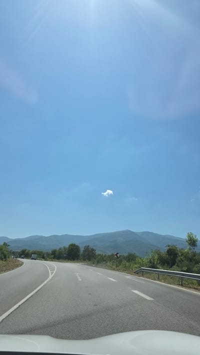 The image depicts a curved asphalt road with white lane markings, leading through a rural, mountainous landscape. The road appears to be two lanes wide, with one lane curving to the left and another to the right. The sky is a clear, bright blue with a few wispy clouds. In the distance, rolling green mountains are visible, covered with trees. There is a guardrail on the right side of the road, and a road sign with a red chevron indicating a curve. The image is taken from the perspective of a vehicle, with the dashboard visible at the bottom. There are no people clearly visible, but there are some vehicles in the distance on the left side of the road. The scene suggests a sunny day, likely during daytime. The location is identified as Mezdreya, Bulgaria.
