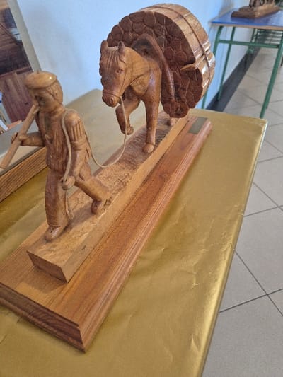 The image displays a carved wooden sculpture depicting a male figure leading an equine animal laden with logs, situated in a room within El Espinar, Spain. The sculpture, crafted from light brown wood, features a man wearing a cap, tunic-like shirt, and trousers, carrying a long stick over his left shoulder. His left hand holds a thin rope, which extends forward to the bridle of a horse or mule. The animal is positioned behind the man, carrying a large, semi-circular stack of individually carved logs secured to its back. Both figures are mounted on an elevated, multi-tiered wooden base. A small, rectangular, brass-colored plaque is affixed to the right side of the base.
The sculpture rests on a surface draped in metallic gold paper or fabric, exhibiting some creases and folds. In the background, a light-colored wall is visible, along with a grey and white tiled floor featuring square patterns. To the right and rear of the sculpture, part of a blue-topped table with prominent green metal legs is present. The overall lighting suggests an indoor setting, likely a public or exhibition space.