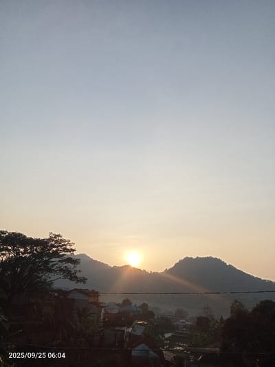 This image depicts a sunrise scene in Ciputri, Indonesia, timestamped 2025/09/25 06:04. The upper two-thirds of the frame show a clear sky, transitioning from a light blue at the zenith to a warm, pale yellow-orange hue near the horizon. The sun, a bright yellow-orange orb, is positioned slightly left of center, emerging from behind the crest of a dark, densely vegetated mountain range, casting visible sunbeams horizontally across the scene. Below the mountains, a residential area is visible, characterized by numerous houses with varied rooflines, appearing in silhouette or dimly lit by the nascent sun. Dark green foliage from trees and bushes is interspersed throughout the foreground and midground, particularly prominent on the far left. A dark, horizontal line, consistent with a utility wire, traverses the lower mid-section of the image, partially obscuring the distant landscape and residences. No individuals are visible.