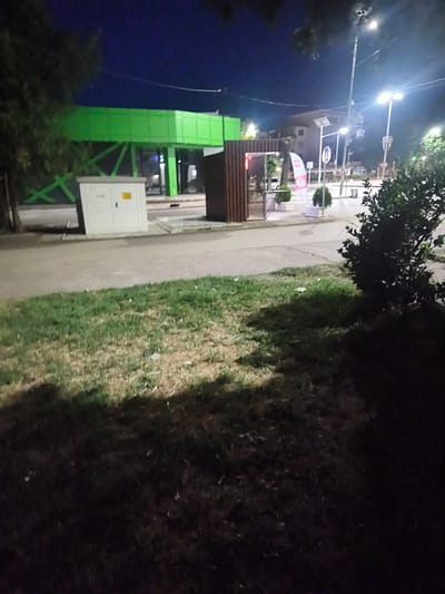 The image depicts a nighttime scene in the city of Harmanli, Bulgaria. In the foreground, a patch of grass is visible, partially illuminated by artificial light sources, casting shadows. Behind the grass, a paved area with a sidewalk runs horizontally across the image. A white electrical box with a yellow warning sticker is positioned on the sidewalk.
Further back, a structure with a slatted wooden exterior and a glass entrance is situated. Beyond this, a bright green, modern-looking overhead structure, possibly a pedestrian bridge or a canopy, is prominent. In the background, several streetlights illuminate the scene, along with buildings and trees. The sky is dark, indicating it is night. There are no people visible in the image.