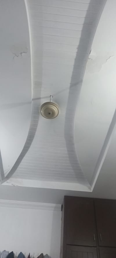 This image shows a domed ceiling with a white, textured surface, possibly made of plastic or painted plaster. The ceiling has a central fan hanging from it. The fan is cream-colored and has a classic design with a round motor housing and a hook for suspension. The ceiling itself has a decorative, curved design that creates a sense of depth and architectural interest. There are some minor imperfections or signs of wear on the ceiling, such as peeling paint or water stains, visible in the upper left and upper right portions of the image.
Below the ceiling, a portion of a dark brown cabinet is visible on the right side. The cabinet appears to be made of wood and has a modern design with two doors, each with a small, thin handle. The lower part of the image shows a white wall with some decorative molding near the top. Below the molding, a rack with several items, possibly clothes or bags, is hanging.
The scene is indoors, suggesting a room in a residential or commercial building. The lighting appears to be artificial, casting subtle shadows on the ceiling. The overall impression is of an interior space, likely a room in a house or an office, given the presence of the ceiling fan and the cabinet. No people are visible in the image. The context provided places the location in Peshawar City Tehsil, Pakistan.