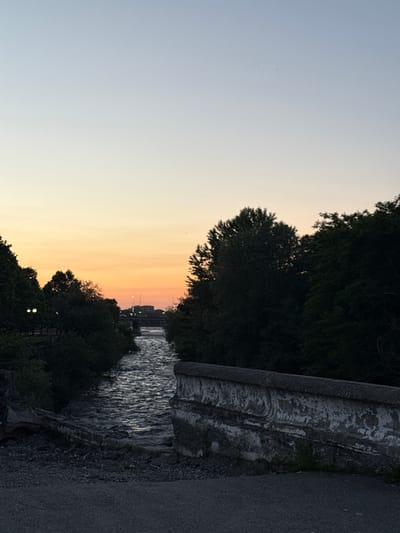 The image depicts a scenic view of a canal or river at dusk. The sky is a gradient of pale blue at the top, transitioning to a warm orange and pink hue near the horizon, indicating sunset. Dark, dense trees line the banks of the water, their silhouettes contrasting with the soft sky. The water itself appears somewhat choppy, reflecting the dim light.
On the right side of the image, a weathered concrete structure runs along the bank, possibly a bridge abutment or retaining wall. Its surface is textured and shows signs of age and wear, with moss or algae visible in some areas. Adjacent to this structure is a paved pathway or road.
In the distance, across the water, a cityscape or town is faintly visible, with lights from buildings and streetlights starting to illuminate. A bridge can also be seen spanning the water in the far distance. There are no people clearly visible in the frame.
The overall mood is peaceful and serene, characteristic of a quiet evening by the water. The time of day is clearly dusk or twilight. The location context is Otf Kaktugay, Russia, though no specific landmarks in the image directly confirm this without external knowledge. There is no visible text in the image.