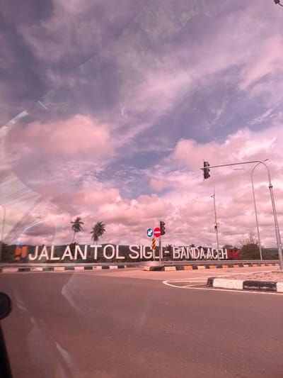 The image shows a street scene with a large sign that reads "JALANTOL SIGLI - BANDA ACEH". The sign is illuminated and appears to be made of metal letters. In the background, there are palm trees and a cloudy sky with pink and purple hues. There are also traffic lights and streetlights visible. The scene is a road with asphalt and white and black striped barriers. The photo appears to be taken from a moving vehicle, as evidenced by the reflections on the surface and the slight blur. The location is identified as Lambadalhok, Indonesia.