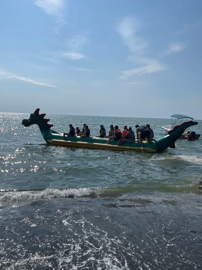 The image shows an inflatable dragon-shaped boat on the water. Several people are seated on the boat, holding onto handles. They appear to be enjoying a recreational activity, possibly a water sport. The boat is green with red accents and a yellow underside. A smaller boat with a canopy is visible in the background. The water is calm with some ripples, and the sun is shining, creating reflections on the surface. The sky is clear with a few wispy clouds. The overall scene suggests a sunny day at a beach or lake, likely in a tourist area given the recreational nature of the activity. The orientation of the image is portrait, with the boat running vertically from bottom to top. The perspective is from a slightly elevated viewpoint, looking down onto the boat and the water.