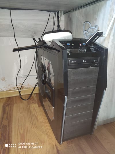 This is a forensic, matter-of-fact description of a computer tower and its surroundings. The computer tower is black and is positioned under a wooden desk. It has a visible optical drive with "M-DISC" and "LG" branding. Several USB ports are visible on the front of the tower, labeled USB1, USB2, USB3, and USB4. A white wireless adapter with an antenna is plugged into one of the USB ports. Black cables are routed behind the computer tower and are managed with zip ties. A white device, possibly a router or modem, is also visible on top of the computer tower. The desk has a light wood grain finish. The wall behind the computer shows some staining. The floor is made of light-colored wood planks. The image also contains a watermark in the bottom left corner that reads "SHOT ON MI 9T AI TRIPLE CAMERA". The location is stated to be in the city of Khazar Raion, Azerbaijan.
