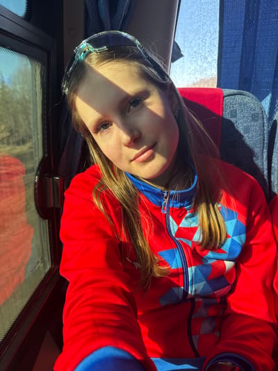 A young fair-skinned girl with light brown hair, wearing clear goggles on her forehead and a red and blue zip-up hoodie, lies at an angle. A bright light casts a shadow across her face. She appears to be inside a vehicle; a bus seat is visible in the background, and a partially visible window is located at the bottom of the frame. It is not possible to confirm her location is Челва, Russia based on visual information.