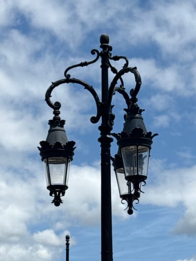 This is a close-up, vertical, eye-level shot of a black ornate lamppost against a bright blue sky filled with white, fluffy clouds. The lamppost has three glass-enclosed lights, two on one side and one on the opposite side, branching out from a central pole. The lamppost's decorative elements are intricate and detailed, with scrollwork and finials. A smaller, single-bulb lamppost is visible in the distance below the main lamppost. The image appears to be taken in Bordeaux, France, likely on a street with historical architecture given the lamppost's design.