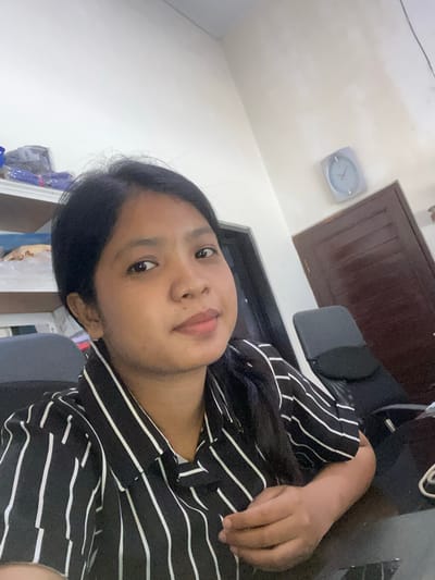 A woman with dark hair is photographed indoors. She is wearing a black and white striped shirt. Behind her, there is a door and a clock on the wall. The location is determined to be Negara, Indonesia.