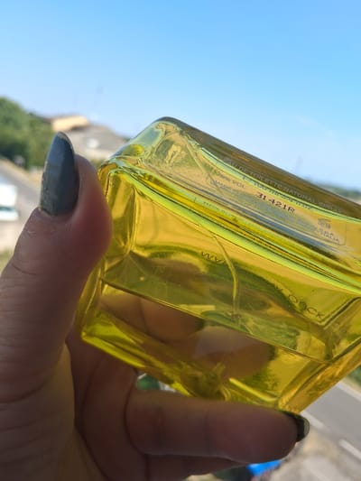 A close-up shot shows a person's hand holding a yellow glass perfume bottle against a bright blue sky. The bottle appears to be a natural spray Eau de Toilette, with text on its surface indicating a volume of 100 ml or 3.4 fluid ounces, and a product code of "31421R". Other visible text includes "79% VOL.", "MADE IN IT" (likely Italy), "Mini Beauty S.p.A.", "Milano - ITALIA", and the brand name "MONCK" or similar. The bottle is tilted, and the sunlight reflects off its glass surface, creating a bright, summery atmosphere. The background suggests an outdoor setting, possibly a balcony or window, with some blurred greenery and buildings visible below. The person's fingernails are painted a dark, metallic color. The image has a natural, candid feel, likely taken during daylight.