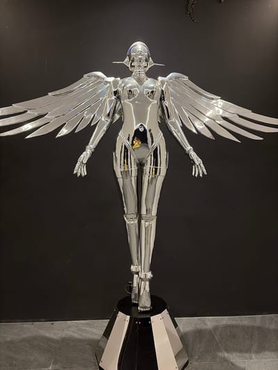 A full-shot, eye-level, studio photograph captures a striking, life-sized sculpture of a chrome-plated humanoid figure with large, metallic wings, set against a plain black background. The sculpture stands on a multi-faceted, dark pedestal with silver accents, and the overall lighting emphasizes its reflective, polished surface. Two small, dark, lizard-like figures are visible on the upper part of the black background, adding an unexpected detail to the scene. The sculpture's features are stylized, suggesting a robotic or angelic aesthetic. The floor is tiled with large, dark gray, rectangular tiles. The scene does not depict any specific location in Baan Lipa Noi, Thailand, or any discernible outdoor environment.