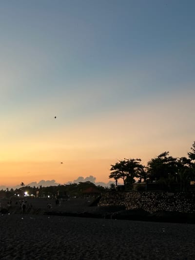 The image captures a sunset view at a beach in Pererenan, Indonesia. The sky transitions from blue to orange as the sun sets. Silhouetted figures of people are visible on the beach near the waterline. Trees and a rock wall line the edge of the beach. Kites are flying in the sky.
