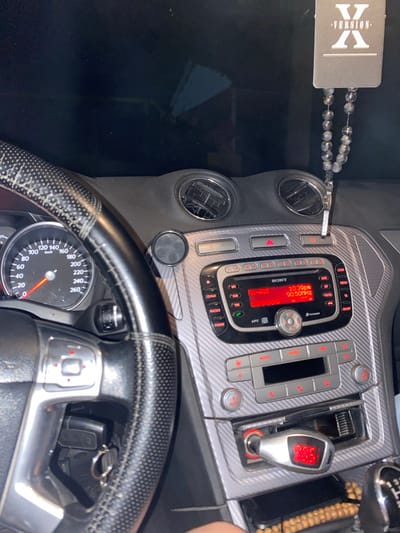 The image shows the interior of a car, specifically the dashboard and steering wheel. The dashboard features a Sony car stereo with a red digital display showing "10:39PM" and "90.50MHz". There are also air vents, various buttons, and a cigarette lighter adapter with a digital display showing "00.0". The steering wheel is wrapped in black leather with white stitching. A black car air freshener with "VERSION X" written on it and a string of beads hangs from the rearview mirror. The car appears to be parked, as indicated by the stationary speedometer reading. The scene is indoors, likely within a garage or on a street in the city of Strășeni, Moldova, based on the provided context. No individuals are visible in the image.