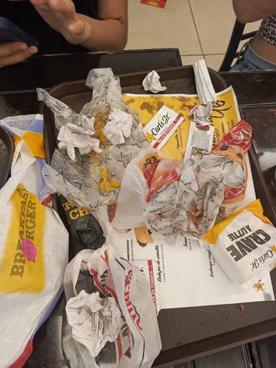 The image displays a cluttered brown fast-food tray filled with discarded wrappers, paper, and used condiment packets, typical of a meal recently finished at a Carl's Jr. restaurant in Samborondón, Ecuador. In the upper left, a person's hands are visible, holding a blue smartphone, while another person's lower torso and leg in blue jeans are partially seen in the upper right background.

The tray is predominantly covered with crumpled white paper wrappers, some with food stains, and a few plastic baggies. Several small, wadded-up paper napkins are also scattered among the trash. Prominently featured are multiple wrappers and a paper bag with the "Carl's Jr." logo and branding. Various text excerpts are visible on the wrappers and packets, including "BREAKFAST BURGER" and "CRAVE A LITTLE." Condiment packets are also present, marked "SALSA DE TOMATE" (Tomato Sauce) and "LOS ANDES." Other legible text includes ingredient descriptions such as "Rodajas de cebolla Cortadas a mano en nuestra cocina." (Onion slices hand-cut in our kitchen) and "Lechuga cruji Crunch" (Crispy lettuce crunch). The scene depicts the aftermath of a fast-food meal, with the items awaiting disposal.