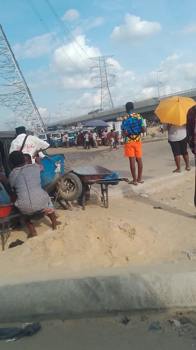 The image captures a street scene in Effurun, Nigeria, under a partly cloudy sky with visible power lines and a bridge in the background. Several people are present; one individual stands out in bright orange shorts and a colorful shirt. Another person is holding a yellow umbrella. In the foreground, there's a makeshift cart or vehicle with people sitting and interacting around it. The ground is covered with sand and debris. There are also a few auto rickshaws in the vicinity.