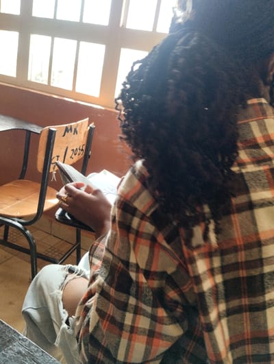 A person sits at a desk near a window. The person is wearing a flannel shirt and ripped jeans, holding a book or notebook. The chair next to them is light brown with a black frame. Inscribed on the chair is the abbreviation "MK" over the date "CT/2055." The window has multiple panes.