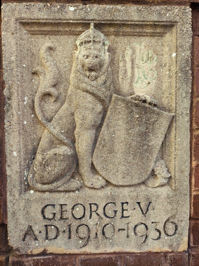 The image shows a stone relief carving of a crowned lion holding a shield, with text below it. The text reads "GEORGE V" and "AD 1910-1930". The carving is set into a reddish-brown brick wall. The style of the carving and the inscription suggest it is a memorial to King George V, likely from the period of his reign or shortly after. The stone is weathered and shows signs of age. The location is specified as being in the city of Maghull, United Kingdom. There are no people or animals present in the scene other than the carved depiction. The environment is an outdoor setting with a brick wall as the backdrop.