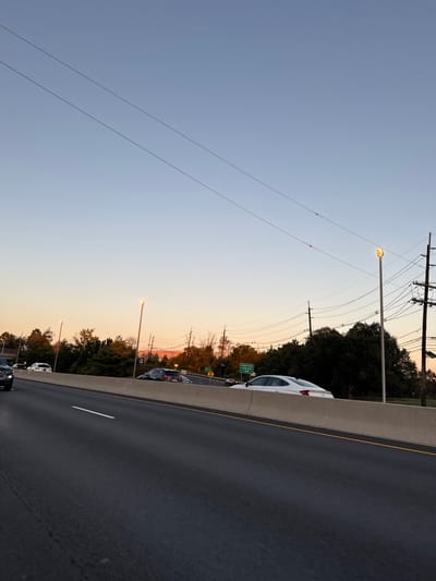 The image depicts a multi-lane highway scene at dusk or dawn in West Windsor, New Jersey, United States, indicated by the sky's blue-to-orange gradient and active streetlights. The foreground features a dark asphalt road with visible white lane markings. A light gray concrete median barrier separates the primary roadway from an adjacent road or embankment to the right.
Along the right side, behind the barrier, multiple vehicles are present. A white sedan is visible partially obscured by the barrier, followed by a dark grey SUV/crossover further back on what appears to be an exit or service road. A green directional sign with white lettering, reading "Carnegie Center," is visible behind the barrier. Active streetlights, emitting a yellowish glow, are mounted on tall poles along the road. Beyond these, a dense line of trees with varied autumnal foliage is visible. Numerous utility poles with multiple power lines crisscross the upper right portion of the image against the fading light. Two prominent parallel power lines span the upper sky. A faint crescent moon is discernible in the upper right quadrant of the sky.
