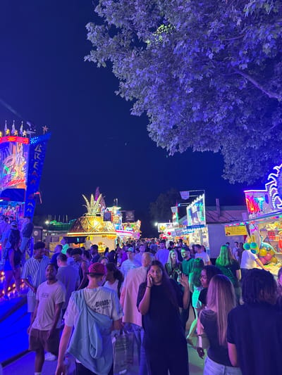 The image depicts a night scene at what appears to be a fair or carnival, likely in Kaiserslautern, Germany. The foreground is filled with people, many of whom are looking at the camera. There are various fair rides and game booths visible in the background, illuminated by bright, colorful lights. A tree with leaves lit in blue is present on the left side. There is a general sense of activity and vibrancy in the scene.