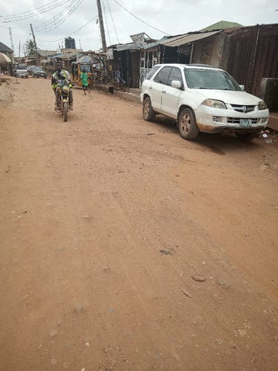 The image shows a dirt road in Ota, Nigeria, located at 6.684° N, 3.178° E. A white SUV, with the license plate “BG 593 AY,” is parked on the right side of the road, with a man riding a motorcycle in the center of the frame. The road is unpaved, and there are buildings on the right side of the road. Several power lines run overhead. The environment is rural and dusty. The man is wearing a green shirt and a blue helmet. The SUV is clean and has its windows up. The buildings on the right are simple, with corrugated metal roofs. The sky is cloudy. The scene is empty of other people except for the man on the motorcycle.