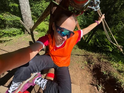 This is a first-person perspective photograph taken outdoors, likely on a sunny day given the strong shadows. The scene appears to be a forest or wooded area, with trees and vegetation visible.
The primary subject is a person, seen from the waist up, lying on their back with their head angled upwards towards the camera. They are wearing an orange vest over a red and white shirt, black pants, and distinctive black and white patterned socks with pink athletic shoes. They are also wearing large, mirrored sunglasses. A purple wristband is visible on their left wrist.
The person's arms are extended, with their left hand holding onto a bundle of thick ropes. These ropes, along with other lighter green and blue ropes, are attached to a wooden structure which appears to be part of some kind of adventure course or rigging. The person's legs are also visible, with one leg bent and the other extended. Red tape is wrapped around what appears to be a wooden support beam near their legs.
The background shows the earthy ground, grass, and the trunks and foliage of trees. The overall impression is of someone participating in an outdoor activity involving ropes and heights, possibly zip-lining, climbing, or a similar adventure sport. The lighting suggests it is daytime. There is no visible text in the image.