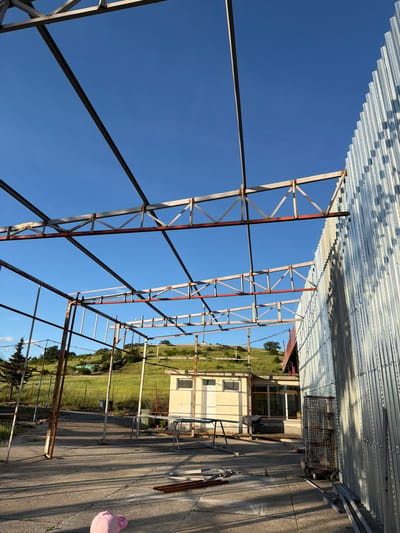 The image shows a partially constructed metal framework against a clear blue sky in Dabnik, Bulgaria. The framework has a rusted appearance in sections. Above the framework is corrugated metal roofing. Beyond the framework, a grassy landscape slopes upward towards a building with light-colored walls and visible windows. On the paved ground adjacent to the framework, there is a metal shopping cart, metal pipes, and a pink object. The image is oriented such that the sky appears vertically in the middle.