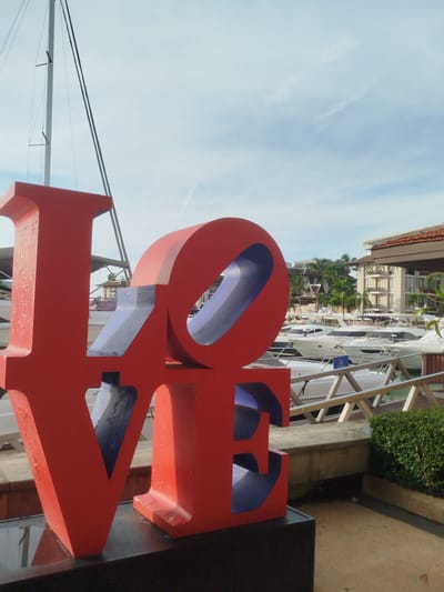A large red sculpture spelling out "LOVE" with blue interior cutouts is positioned in the foreground of the image. The sculpture is placed on a dark base, in front of a marina. In the background, numerous white yachts are docked. Beyond the marina, buildings with tiled roofs and balconies are visible, interspersed with palm trees, suggesting a tropical environment. The sky above is partly cloudy with a light blue hue. A sailboat mast extends upwards on the left side of the frame. The scene is located at the Boat Lagoon Phuket, Ko Kaeo, Thailand.