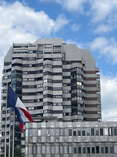 This media file captures a striking perspective of modern architecture under a dynamic sky. The primary subjects are two adjacent multi-story buildings, characterized by their geometric, somewhat brutalist designs. The building on the left features a facade with alternating bands of grey concrete and windows that appear to be set at an angle, creating a visually complex, almost undulating surface. The building on the right presents a more uniform facade of reflective metal panels and rows of rectangular windows, giving it a sleek, modern appearance.
In the foreground, the lower portion of a flag is visible, displaying the colors of the French flag – red, white, and blue. The flag appears to be waving, suggesting an outdoor setting and possibly a breezy day. The sky above is a vibrant blue, dotted with fluffy white clouds, indicating it is daytime, likely mid-morning or afternoon.
The scene is set against a backdrop of a cloudy blue sky, with the buildings dominating the frame. The unusual angles and repetitive patterns of the architecture suggest an urban or suburban environment, likely a city or a business district. There are no people visible in the frame. The overall impression is one of a modern, structured environment, possibly a commercial or residential complex, with a hint of national pride represented by the flag. The weather appears pleasant, with a mix of sunshine and clouds. The architectural style might evoke a sense of the late 20th century. The location context provided is Créteil, France, which aligns with the presence of the French flag.