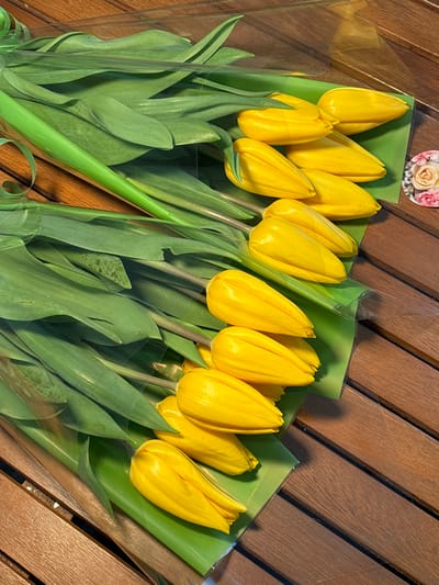 The image displays two bouquets of fresh yellow tulips arranged diagonally on a dark brown wooden surface. Each bouquet contains approximately five to six closed or partially closed yellow tulip buds with vibrant green stems and leaves. The tulips are individually wrapped in clear plastic film, beneath which a layer of light green paper or cellophane is visible. A thin green ribbon is tied around the upper portion of the clear wrapping of the top bouquet. The wooden surface exhibits distinct plank lines and grooves. In the upper right corner, a small, round decorative sticker featuring an image of pink and cream roses is partially visible. No individuals are present in the frame. The specific location in Slavyansk-na-Kubani, Russia, is not discernible from the visual contents of the image.