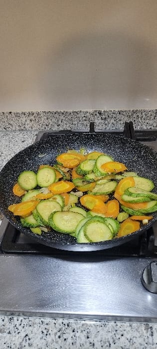 The image displays an overhead view of a black, speckled frying pan filled with an assortment of sliced vegetables, positioned on a stovetop. The pan contains numerous circular slices of green zucchini and crescent-shaped or oval slices of orange carrots, mixed with what appears to be finely diced white onion. The vegetables are lightly seasoned with visible specks of black pepper.

The pan is situated on a black metal burner grate, indicating it's placed on a stovetop. To the right of the pan, a brushed stainless steel surface, likely part of the stove, is visible, along with a control knob at the upper right edge of the frame. A light-colored, speckled countertop, possibly granite, surrounds the stovetop area. To the left, a plain, light beige or tan wall forms the background. The overall scene depicts a culinary activity, specifically the cooking or preparation of vegetables in a kitchen environment. No people are visible, and there is no visible text. The setting is consistent with a domestic kitchen in Tixeraïne, Algeria.