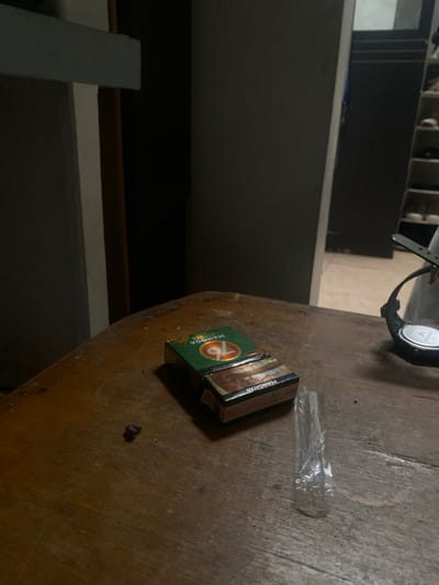 The scene is an indoor environment in Cimenyan, Indonesia, depicted from a slightly elevated perspective overlooking a wooden table. The table surface, exhibiting a worn appearance with visible grain and minor abrasions, holds several items. In the immediate foreground, a small, dark, irregularly shaped object, consistent with a piece of debris or tobacco, is positioned to the left. Central on the table is a green and brown pack of "76 Mangga" cigarettes, with its top flap partially open, revealing contents within. Adjacent to the cigarette pack on the right is a crumpled, transparent plastic wrapper. Further to the right, a black digital wristwatch with a circular metallic face and a black band rests on its side.
The background reveals the interior space, which is dimly lit. On the right, a dark, vertical structure, identifiable as a wardrobe or shelving unit, displays multiple internal compartments, some appearing to contain various items like shoes or folded textiles. A lighter-colored floor is discernible beyond this structure. On the left, a light-toned vertical partition or door frame is present, leading into a darker, undefined space. No individuals are present, and no direct actions or interactions are depicted within the frame; the scene is static.