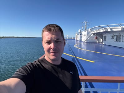 A man is taking a selfie on the deck of a ferry. He is wearing a black t-shirt and is smiling at the camera. The ferry deck is painted blue with yellow markings. In the background, there is the ocean, a distant island, and the upper structures of the ferry, including a helideck and a navigation tower. The sky is clear and blue. The scene appears to be in the waters near Karlskrona, Sweden, given the presence of ferries and the archipelago typical of the region.