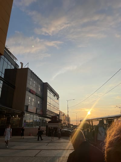 The image depicts a street scene at sunset in the city of Dnipro, Russia. In the foreground, several people are walking on a paved pedestrian area. To the left, a modern building with a glass facade and upper floors featuring multiple windows dominates the scene. Red lettering on the building reads "АПТЕКА" (Apteka), indicating a pharmacy. Further to the right, a bus stop shelter is visible, with people waiting. The setting sun casts a warm glow, creating lens flares and illuminating the sky with a gradient of orange, yellow, and blue, interspersed with scattered clouds. Power lines crisscross the upper portion of the image. A few cars are parked or moving on the street in the background.