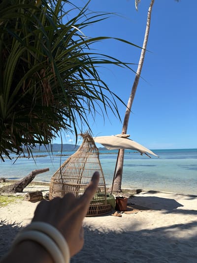 A person's left hand, adorned with two white bracelets, is in the foreground, with the index finger pointing towards the right. Beyond the hand, on a sandy beach, is a large, woven, egg-shaped chair with green cushions. Behind the chair, a thin, tall palm tree stands next to a partially open, light-colored umbrella supported by a wooden pole. The ocean, with clear, turquoise water, stretches to the horizon, where a line of hills and a distant boat are visible under a bright blue sky. To the left of the frame, large, dark green leaves of a tropical plant hang from above, casting shadows on the scene. The location is Baan Thong Krut, Thailand.