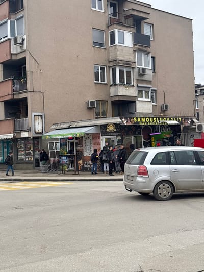The image depicts a street scene in Vitez, Bosnia and Herzegovina, specifically focusing on the ground level of a multi-story, light brown apartment building. The building features various windows, some with external shutters, and multiple balconies, some enclosed, along with several visible air conditioning units.
At the street level, there are multiple commercial establishments. To the left, a store with a turquoise awning is visible. Adjacent to it, a kiosk with a green and white striped awning displays "STOLNI FUDBAL MOGOMET" (table football) and advertising for "TOPLA ČOKOLADA" (hot chocolate) and coffee. Directly to its right is another self-service kiosk with a dark awning featuring "SAMOUSLUGA 24/7" (self-service 24/7) and "KAVA GRICKALICE ZA PONJETI" (coffee snacks to go) signage, illuminated by a purple neon light.
On the asphalt road in the foreground, a silver Opel Zafira station wagon is positioned with its rear facing the viewer. The vehicle bears a Bosnian license plate, M34-K-602, and has a visible tow hitch. A pedestrian crossing, marked with yellow paint and white stripes, is visible on the street to the left of the car.
Multiple individuals are present. On the sidewalk to the far left, a male pedestrian wearing a dark jacket and jeans walks from left to right. A small group of people is visible near the kiosk with the green awning, including one individual seated at an outdoor table. A larger group of approximately 8-10 young males, predominantly wearing dark jackets and backpacks, are congregated directly in front of the "SAMOUSLUGA 24/7" kiosk, potentially queuing. Another adult male in a lighter jacket is visible to their right near the same kiosk. The overall scene suggests a typical urban environment during daylight under an overcast sky.