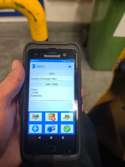 A person's hand holds a Honeywell handheld scanner in Komorniki, Poland. The scanner displays an application interface showing shipment details. The screen indicates "01 SSCC" and a barcode with the number "542501272233011491." Below this, it lists "KRT / NOS" and "Palet: 1 EUR Kartonów." There are several icons at the bottom of the screen, including "EUR," a stack of boxes with a red "X," "H1," a left arrow, a blue truck, and a green checkmark. The background shows a yellow industrial railing and a green waste bin.