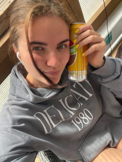 A young woman with blonde hair, blue eyes, and light skin is taking a selfie. She is wearing a grey hooded sweatshirt with the words "DELICATE 1980" printed in white on it. She is also wearing white earbuds. In her right hand, she is holding a yellow can of what appears to be a beverage. The background is blurred but suggests an outdoor setting with wooden structures and some sort of ribbed material. There are no identifiable city landmarks or specific locations within Мосметаллоконструкция Онт, Russia visible in the image.