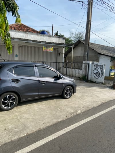 A grey Honda Brio is parked on the right side of a paved road in Purwakarta, Indonesia, with a white stripe separating the road from the sidewalk. The car is facing towards the left, and its windows are tinted. Behind the car are a set of black metal gates and a house with a white wall and a yellow sign reading "DI KONTRAKKAN HAKIMI 0817 7080 2113" in front of it. The sidewalk is also paved with concrete. To the right, there is a second house with a grey roof and a white wall with a large graffiti image of a red, white, and black cartoon face. There is also a yellow sign on the white wall that is partially visible. There is a small amount of green vegetation in the background. The sky is light blue with thin white clouds.