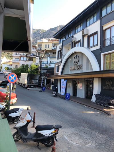 In Kaş, Turkey, on a sunny day, a street scene unfolds with several vehicles and businesses. In the foreground, two scooters are parked on the sidewalk. One white scooter is positioned upright, while another white scooter is parked further back. A red car with its trunk open is also visible. A large delivery truck with its back doors open and boxes visible inside is parked on the street. A man in a light-colored shirt stands near the truck. To the right of the truck is a traffic sign with a blue circle and a red cross.

Dominating the right side of the image is a modern building with a large arched entrance to a store labeled "MUHTAR". Above the entrance, the "MUHTAR" logo is prominently displayed. Inside the store, shelves are visible. Multiple shopping carts are lined up outside the store entrance. Banners and signs are displayed in front of the store, some with Turkish text.

In the background, other buildings, including a hotel, are visible, with apartments and windows. Air conditioning units are mounted on some of the buildings. A power pole with electrical equipment stands in the middle ground. Mountains are visible in the distance, indicating the hilly terrain of the region. The street is paved with interlocking stones.