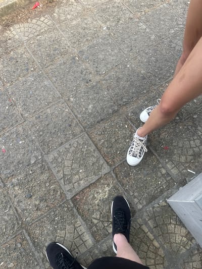 This media file shows a close-up, overhead view of two people's legs and feet standing on a textured concrete surface. The concrete is laid in square and semi-circular tiles, with visible grout lines and scattered debris, appearing to be twigs and small stones.
One person is wearing white canvas sneakers with a black and white logo print, which appears to be the "Christian Dior" logo repeated. This person's legs are smooth and tanned, with one visible knee.
The other person is wearing black athletic shoes with white soles and reflective laces. Only the lower legs and feet are visible, with one leg clad in black fabric, likely pants or shorts.
The scene suggests an outdoor setting, possibly a public walkway or plaza, given the paving. The lighting indicates it is daytime, and the debris on the ground suggests an area that is not meticulously maintained, common in outdoor public spaces. There is no indication of specific weather conditions beyond the presence of daylight. The image focuses on footwear and legs, with no faces or explicit emotional cues visible. The overall impression is casual, possibly a moment captured while walking or pausing.
The provided location context is Ovoshtnik, Bulgaria.