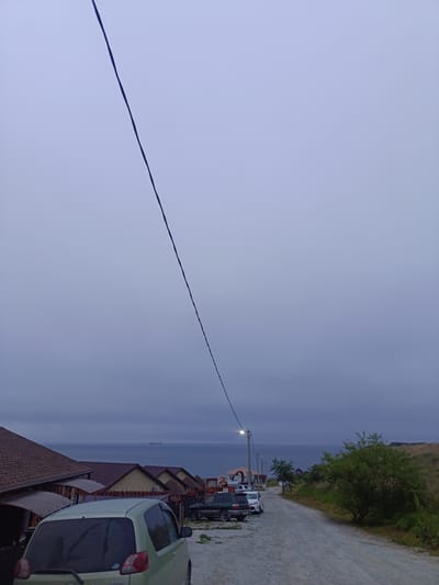 The image captures a view of a street leading towards the sea in Zarubino, Russia. The sky is overcast and a utility wire stretches diagonally across the frame from the top left to the bottom center. On the left side of the street, a row of houses with reddish-brown roofs are visible. In front of the houses, several cars are parked. A light-colored minivan is parked in the foreground on the left. Further down the street, a dark green SUV and a white car are parked. On the right side of the street, a dirt road curves upwards, with a grassy hill and green bushes on the far right. The sea is visible in the background, with a ship on the horizon. The overall atmosphere is subdued due to the cloudy weather.