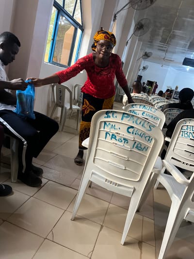 A woman in a red lace blouse and colorful wrap exchanges a blue plastic bag with a seated person. Rows of white plastic chairs, labeled "St. Paul's MIL. Church, Proto IBN", stack behind them. The setting appears to be inside a church, with ceiling fans and a window visible. The tiled floor reflects the interior light, adding to the brightness of the space.