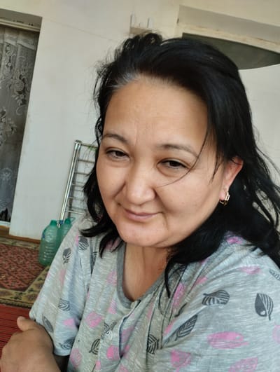 A middle-aged woman with black hair and fair skin is photographed from the chest up in an indoor setting. She is wearing a gray, short-sleeved shirt with a repeating pattern of pink and black leaves. A gold hoop earring is visible in her right ear. The woman is looking towards the camera with a slight smile and her head tilted. In the background, to the left of the woman, there is a metal drying rack with two green plastic bottles. The wall behind her is a light color, and a patterned rug is visible on the floor. This image appears to have been taken in a residential area of Yangibazar, Uzbekistan.