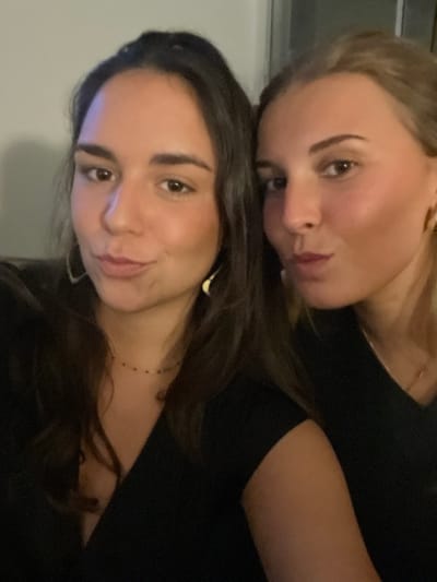 This image depicts two young adult females captured in a close-up, chest-up selfie within an indistinct indoor setting.
The woman on the left has long, dark brown hair and brown eyes. She looks directly towards the camera with a subtle smile. She wears a dark, V-neck top, a thin gold necklace with small dark beads, and prominent light-colored, crescent-shaped earrings.
The woman on the right has lighter-colored hair, styled away from her face, and is positioned closely beside the woman on the left. She is making a puckered, 'kissing' expression towards the camera. She wears a dark, V-neck top, small gold hoop earrings, and a thin gold chain is partially visible around her neck.
Both individuals exhibit warm skin tones. The background is a plain, unidentifiable wall or surface, suggesting an interior space illuminated by soft, artificial lighting. The image provides no visual indicators to confirm a specific location within the city of Bath, United Kingdom.