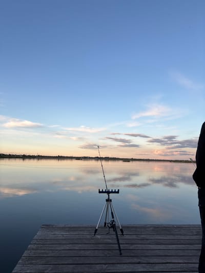 A horizontal view of the sky and water in Khryashchevka, Russia, taken from a low angle on a dock. The sky is visible on the left, transitioning from blue to a lighter, cloud-filled area above the horizon. The water reflects the sky and clouds, creating a mirrored effect. A fishing rod rests on a tripod on the right, positioned on a wooden dock. The dock's boards run parallel to the camera's perspective. A partial view of a person's legs is visible at the top center of the image.
