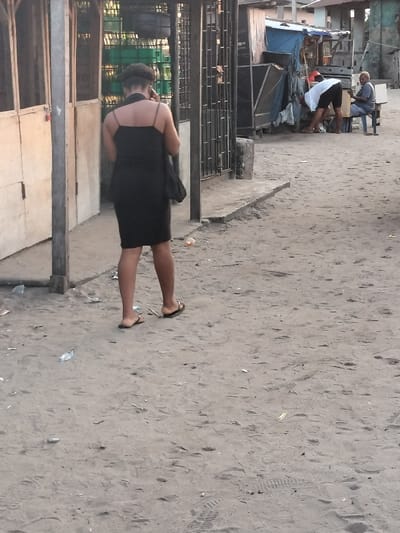 A woman in a black dress and sandals walks away on a sandy road, holding a phone to her ear. Trash litters the ground. Two men stand near a makeshift structure, with one bending over and the other seated. Buildings are visible in the background. The overall scene appears to be a street or alleyway in a less developed area.